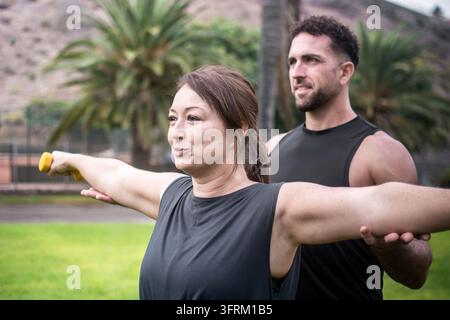 Male personal trainer supporting a woman's arm while she performs a strength training exercise with yellow dumbbells in an outdoor park. Stock Photo