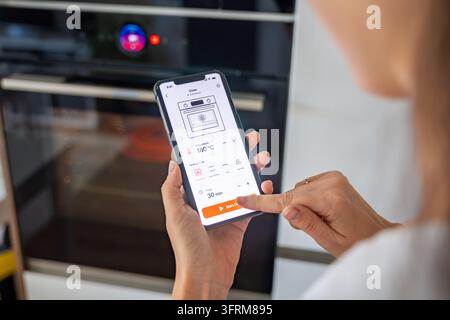 Woman starts a smart oven by tapping a control button on her smartphone in a modern kitchen. Connected home technology enables remote cooking Stock Photo