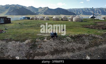 Base camp vibes: two travellers relax on the grass in front of a yurt camp near Kel-Suu Lake, Kyrgyzstan Stock Photo