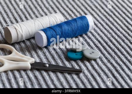 Sewing supplies including spools of white and blue thread, colorful buttons, and scissors on ribbed grey fabric Stock Photo