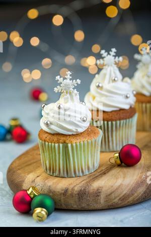 Cupcakes with vanilla cream, snowflakes and colorful Christmas balls on a wooden serving board on a blurred background of lights, selective focus Stock Photo