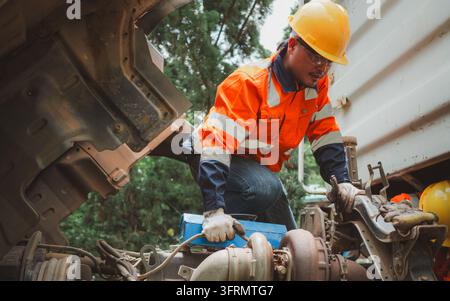 Professional Asian male mechanic in safety helmet and reflective jacket inspecting heavy duty truck engine during vehicle maintenance and repair servi Stock Photo