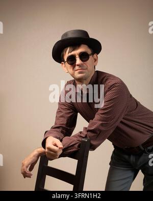 Portrait of young smiling flirtatious man in black hat and black glasses leaning his arm on back of chair. on light studio background Stock Photo