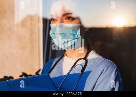Young sad female caucasian UK US NHS-EMS doctor taking care through ICU window, fear insecurity in eyes, wearing face mask gazing at sun, Hope faith i Stock Photo