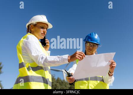 Two women engineers in safety helmets and high-visibility vests review project data beside a wind turbine, coordinating renewable energy work under a Stock Photo