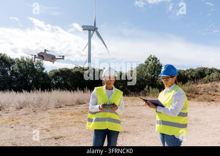 Two female engineers conduct an aerial drone inspection of a wind turbine at a renewable energy site, using remote controls and a tablet while wearing Stock Photo