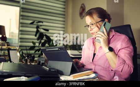 Candid photo of a professional businesswoman in her late 40s working at her desk in an office, speaking on a smartphone while interacting with a table Stock Photo