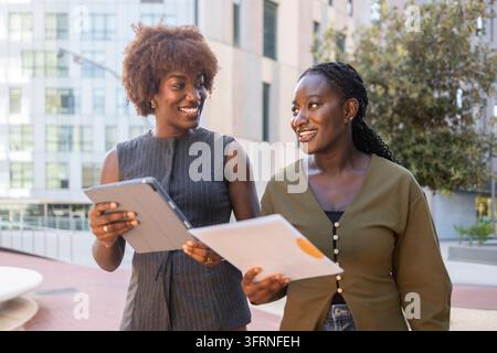 Two smiling Black businesswomen walk outside a modern office building, holding a tablet and documents while discussing work and collaborating on a bus Stock Photo
