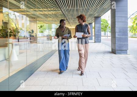 Two Black businesswomen walk beside a modern office building, smiling and discussing documents during a professional meeting in an urban business dist Stock Photo
