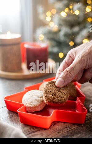 Close-up of a hand taking a traditional Spanish polvoron cookie from a red Christmas tree shaped dish, surrounded by assorted shortbread, candles and Stock Photo