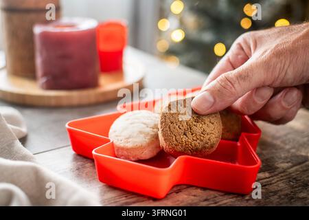 Close-up of a hand taking a traditional Spanish polvoron cookie from a red Christmas tree shaped dish, surrounded by assorted shortbread, candles and Stock Photo