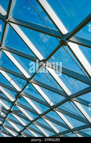 Upward view of a geometric glass roof with metal beams, cables, and structural joints forming an abstract architectural pattern against a vivid blue s Stock Photo