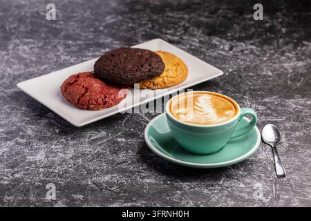Fresh cappuccino with latte art in a mint green cup and saucer, served beside assorted chocolate, red velvet, and oatmeal cookies on a white plate. Stock Photo
