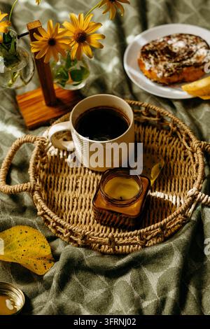 Cozy autumn coffee still life featuring a cup of black coffee on a woven tray, golden flowers, a cinnamon roll, candle, and fallen leaf on textured fa Stock Photo