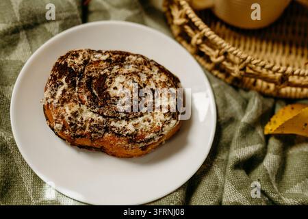 Close-up of a freshly baked cinnamon roll dusted with sugar on a white plate, styled with coffee, a woven basket, green linen, and autumn leaves. Stock Photo