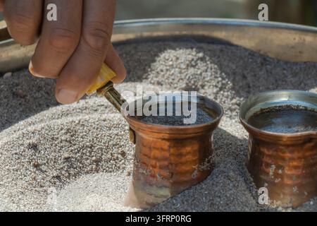 Close-up of a hand holding a copper cezve while traditional Turkish coffee brews in hot sand beside a second pot, showing an authentic manual preparat Stock Photo