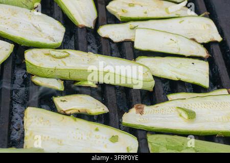 Close-up of lengthwise zucchini slices cooking on a hot barbecue grate, showing fresh green skin, pale flesh and simple outdoor vegetable preparation. Stock Photo