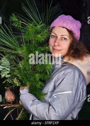 Woman wearing a pink hat is holding a pine tree. She is standing in front of a tree Stock Photo