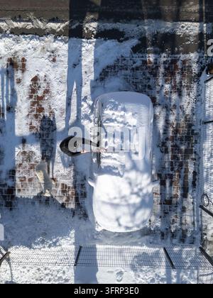 A person is cleaning snow off a car in a residential area during the winter morning. The ground is covered in white snow. Stock Photo