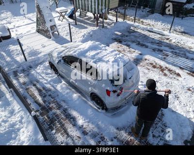 A person is cleaning snow off a car parked in a snowy space during the winter morning. The ground is covered with snow. Stock Photo
