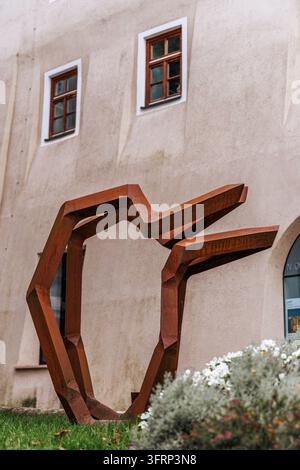 A striking abstract metal sculpture stands in front of a historic building with windows. The sculpture's rusty orange hue contrasts with the building' Stock Photo
