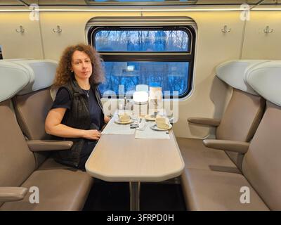 Woman sits at a table in a train car. There are two chairs in the background. A cup and a bowl are on the table in restaurant Stock Photo