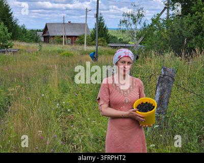 Woman wearing a pink hat is holding a yellow bucket with currant berry. She is standing in a field. There are several houses in village Stock Photo