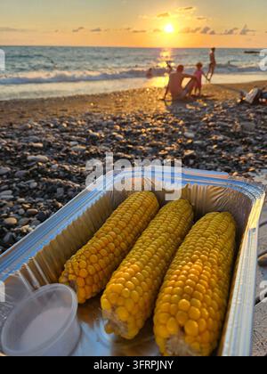 Three yellow corn cobs are in a foil container. The container is on a table. A person is standing behind the container on beach sunset Stock Photo