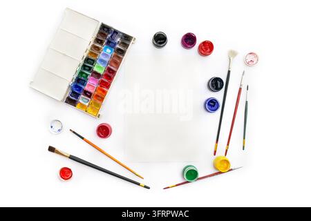 Top-view flat-lay photograph of an open box of watercolor paints, gouache jars, paintbrushes, and a blank sheet of paper on a clean white background. Stock Photo