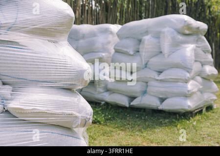 Stacks of white sacks filled with agricultural supplies, wrapped in clear plastic, resting on wooden pallets outdoors for storage Stock Photo