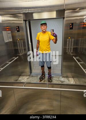 Man in a yellow shirt is taking a picture selfie of himself in an elevator. He is wearing skates and a green hat Stock Photo