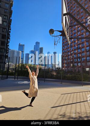 Woman is playing basketball in a park is surrounded by tall buildings. She is wearing a white dress and is jumping up to shoot the ball Stock Photo