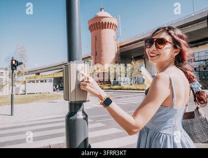 Smiling woman with sunglasses pressing a pedestrian crossing button, waiting to cross a busy street in an urban area Stock Photo