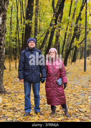 Man and a woman couple middleaged are standing in a autumnal forest. The man is wearing a blue jacket and the woman is wearing a red jacket Stock Photo