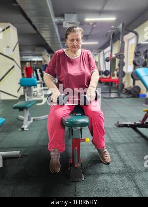 Woman senior in a red shirt and pink pants is holding two dumbbells. She is sitting on a bench Stock Photo
