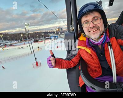 Man in a red jacket is sitting in a chair on a ski slope. He is smiling and giving a thumbs up in city Stock Photo
