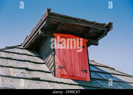 Weathered wood shingle roof with bold red painted attic dormer door set against clear blue sky. Rural heritage concept. Stock Photo