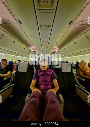 A man in a purple vest is sleeping on an airplane. The man is wearing a mask and has his legs crossed. The other passengers are sitting in the seats around him Stock Photo