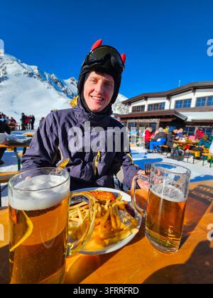 A man is sitting at a table with two glasses of beer and a plate of food. He is wearing a hat and a helmet. The atmosphere seems to be casual and relaxed, as the man is enjoying his meal and drinks Stock Photo