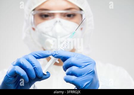 Female NHS medical workers wearing protective suit, goggles, face mask and gloves, holding syringe with needle containing vaccination booster dose, Co Stock Photo