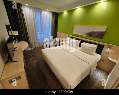 MINSK - MAR 24, 2025: A hotel room with a large bed and a television. The room is clean and well-lit, with a green wall and a green curtain. The bed is neatly made, and there are two pillows on it Stock Photo