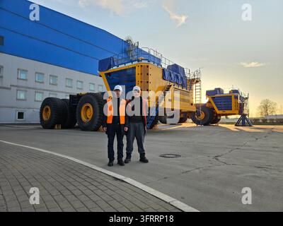Two men standing in front of a large yellow truck. The truck is parked in a lot next to a building Stock Photo