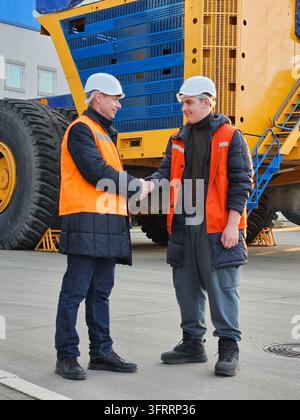 Two men shake hands in front of a large yellow and blue truck. One of them is wearing an orange vest Stock Photo