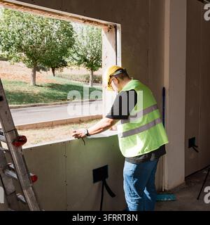 Building surveyor in hard hat and reflective vest measures an unfinished window opening from inside a renovation site, with exterior trees and street Stock Photo