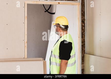 Side view of a building surveyor in hard hat and reflective vest examining an unfinished doorway, drywall panels, insulation and exposed conduits duri Stock Photo