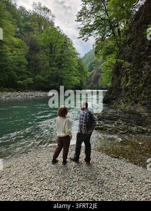 Man and a woman are standing on a rocky shore looking at the water. The man is wearing a backpack Stock Photo
