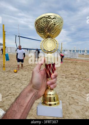 Man holding a gold trophy on beach volleyball game sand - Hand holds golden trophy on sandy beach. People play volleyball in background. Blue sky and clouds complete the scene Stock Photo