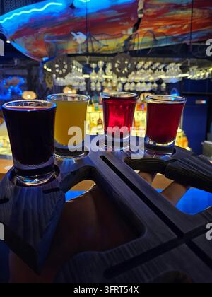 Person is holding a wooden tray with four different colored glasses of liquor. The glasses are arranged in a row, with the first glass being purple, the second orange, the third red Stock Photo