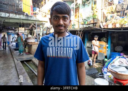 Portrait of a young man in a blue t-shirt standing in a narrow alley of Dhobi Ghat with washing tanks and laundry workers behind him, Mumbai, India Stock Photo