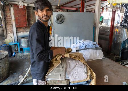 Young man in a black shirt folding trousers and clothes on a table in a laundry workshop with industrial machines at Dhobi Ghat, Mumbai, India Stock Photo